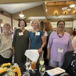 Five women stand and sit, smiling, at a banquet table set with drinks and sunflowers, inside a well-lit event hall with people and a presentation screen in the background.