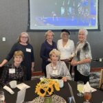 Six women pose together at a round table set for a meal, with a sunflower centerpiece and drinks, in a conference room with a presentation screen in the background.
