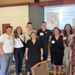 A group of eight women pose together indoors at a professional event; a presentation slide titled "Daytime Bookworms" is visible in the background.