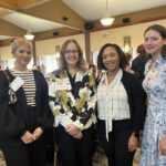 Four women standing indoors at an event, wearing name tags and business casual attire, posing and smiling for a group photo.