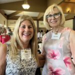 Two women smiling at a social event, both wearing name tags, with a group of people and a presentation screen in the background.