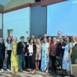 A group of people pose together indoors, some standing and some sitting, with a projection screen and tables visible in the background.