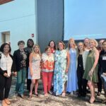 A group of thirteen women stands together indoors, posing for a photo in front of a blank projection screen and light-colored walls.