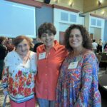 Three women stand together and smile at an indoor event, with other people mingling in the background.