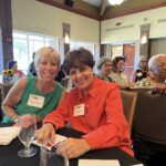 Two women sit at a table smiling at a gathering, with other attendees seated in the background in a well-lit event room.