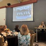 A woman speaks at a podium in front of an audience, presenting a slideshow with a projected group photo labeled "1984-85 Linda’s First Year Coaching - Year Begins.