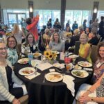 A group of women sit around a table with food and drinks at a banquet event in a large room, with other attendees visible in the background.