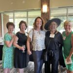 Five women stand together indoors, smiling at the camera. They wear semi-formal attire and nametags, with one woman in a black outfit and hat. Large windows and brick walls are in the background.