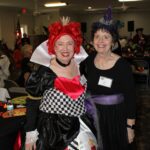 Two women in costumes pose indoors; one dressed as the Queen of Hearts and the other in a black dress with a purple witch hat.