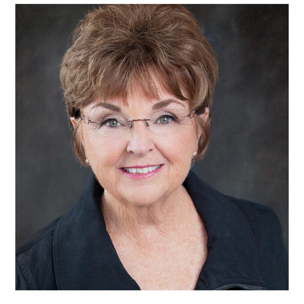 Older woman with short brown hair, glasses, and a black collared top, smiling in front of a dark, neutral background.
