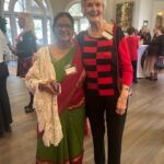 Two women stand together at an indoor event, both wearing name tags. One wears a green and pink sari, the other a black and red sweater. People and tables are visible in the background.
