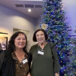 Two women wearing name tags stand in front of a decorated Christmas tree; one holds a plate of food, both are smiling at the camera.