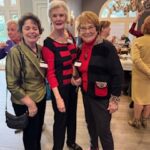 Three older women stand together indoors at a social gathering, smiling at the camera. Other people and a chandelier are visible in the background.