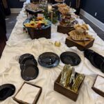 A buffet table with various food items, including fruit, pastries, and finger foods, displayed on platters and stands, with plates and utensils arranged at one end.