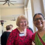Two women wearing name tags smile at the camera at an indoor event, with other people and a ceiling fan visible in the background.