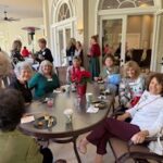 A group of women sit and stand around a large oval table indoors, socializing and eating snacks at a gathering.