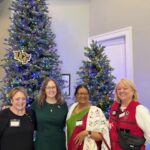 Four women stand smiling in front of decorated Christmas trees with UCF ornaments, wearing festive and formal attire, inside a brightly lit room.