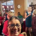 A woman in glasses takes a selfie at a social gathering; several people are mingling in a warmly lit room with food and drinks.