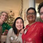 Five women smile for a group selfie indoors with a decorated Christmas tree in the background.