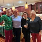 Four women stand together smiling in a kitchen with hanging lights, wearing festive or casual clothing, at what appears to be a holiday gathering.