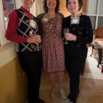 Three women stand together indoors, smiling at the camera; two hold wine glasses and all are wearing name tags.
