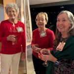 Three older women stand together indoors, smiling and holding drinks. They wear name tags and festive or casual attire, suggesting a social or holiday gathering.