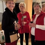 Three older women stand together indoors, each holding a drink and smiling at the camera. They are dressed in festive, winter attire.