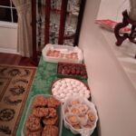 A table covered with a green cloth displays assorted cookies and treats in trays and paper cups, set next to a glass cabinet in a dining room.