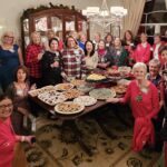 A group of women gathers around a table filled with assorted cookies and desserts in a warmly decorated dining room.