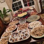 A large dining table is covered with a variety of homemade cookies and desserts arranged on plates and trays in a well-lit dining room.