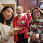 Three women stand together indoors at a gathering; one is holding a plate of food, another wears a red sweater, and the third holds a phone. They are smiling at the camera.