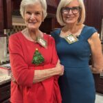 Two older women stand side by side in a kitchen, both wearing name tags; one is in a red sweater with a Christmas tree, the other in a blue dress.