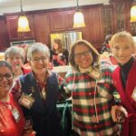 Five women stand and smile in a kitchen during a gathering, each wearing name tags and holding drinks. Other people converse in the background near a kitchen island.