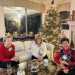 Three women sit on a couch in a decorated living room with a Christmas tree, wrapped gifts, and festive ornaments visible around them.