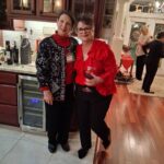 Two women pose together in a kitchen during a gathering. One holds a glass of wine. Bottles and gifts are on the counter, with other people visible in the background.