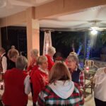 A group of older women gather indoors at night, some talking and others taking photos. The room is decorated with holiday lights and candles.