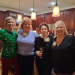 Four women pose together in a kitchen at a gathering, all wearing name tags; one woman is wearing a green Christmas-themed sweater.