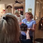 A woman smiling and holding a chocolate birthday cake with lit candles while others stand nearby inside a decorated home.