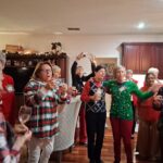 A group of women in festive attire gather in a kitchen, some holding drinks and taking photos, appearing to enjoy a holiday celebration.