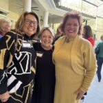 Three women stand closely together, smiling at the camera at an indoor-outdoor social gathering. Two wear name tags and the background shows other people and part of a patio.