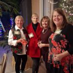 Four women standing together indoors at a holiday gathering, holding drinks and smiling, with festive decorations and a Christmas tree in the background.