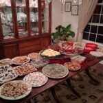 A dining table is covered with various trays and plates of homemade cookies and desserts in a dining room, with a china cabinet and houseplant in the background.