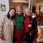 Three women stand together smiling at a holiday gathering, one wearing a green Christmas sweater, with festive decorations and reflections visible in the background.