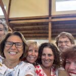Six women smiling and posing for a group selfie indoors, with wooden beams and large windows visible in the background.