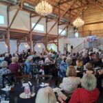 A large group of people sit at round tables in a rustic hall with wooden beams and chandeliers, listening to speakers at the front near a staircase and a presentation screen.