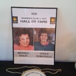 Framed poster reads "2026 Women's Club at UCF Hall of Fame" with photos and names of Merrell Bailey and Sheila Somerville, placed on a table with a decorative plate and beads.