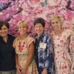 Six women stand together smiling in front of a backdrop featuring pink flowers and butterflies. They are dressed in floral and patterned clothing.