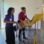 Two people playing steel drums indoors, standing next to a yellow music stand with sheets, both wearing name tags and casual outfits.
