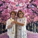 Two older women stand smiling in front of a vibrant backdrop of pink cherry blossoms with butterflies and glowing lights.