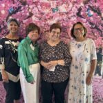 Four women stand side by side, smiling in front of a backdrop with pink cherry blossoms and butterflies.